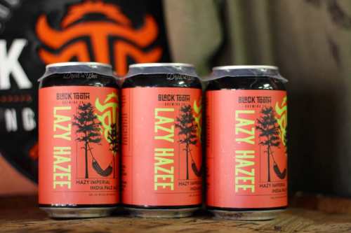 Three cans of "Lazy Hazee" Hazy Imperial India Pale Ale on a wooden surface, featuring vibrant orange and green labels.