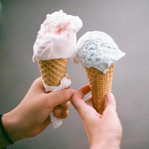 Two hands hold ice cream cones: one with pink cotton candy flavor and the other with light blue mint flavor.