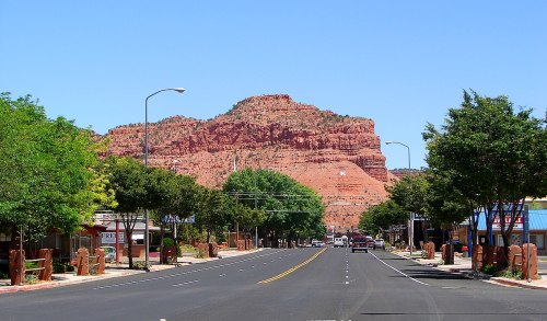 A wide street lined with trees, leading to a red rock formation under a clear blue sky.