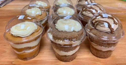 A variety of layered desserts in clear cups, topped with swirls of frosting, displayed on a wooden surface.