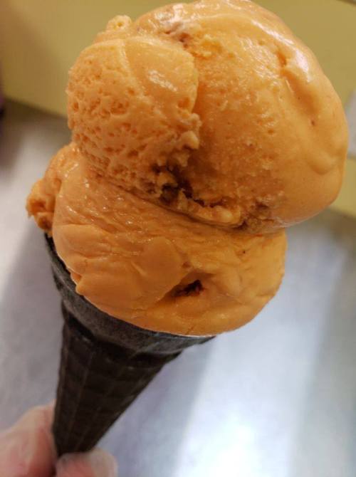 A close-up of a scoop of orange ice cream on a black waffle cone, held by a hand.