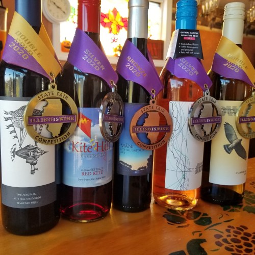 Five bottles of wine with medals and ribbons from the Illinois State Fair Competition, showcasing various awards.