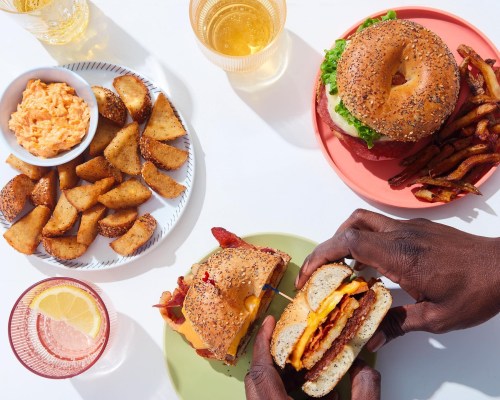 A table spread with a burger, fries, potato wedges, and a bowl of dip, with drinks in the background. Hands holding a burger.