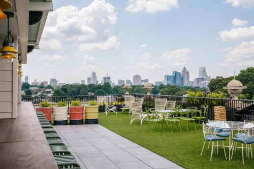 Rooftop view featuring green lawn, white chairs, colorful planters, and a skyline of tall buildings under a blue sky.