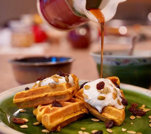 A close-up of waffles topped with yogurt, seeds, and dried fruit, with syrup being poured over them on a green plate.