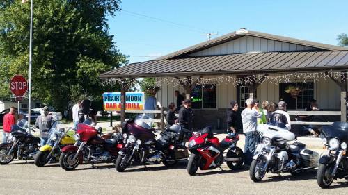 A line of motorcycles parked outside a bar and grill, with people gathered near the entrance on a sunny day.