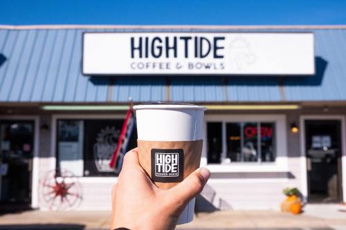 A hand holds a coffee cup in front of a café named "Hightide Coffee & Bowls" on a sunny day.