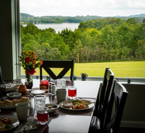 A dining table set with breakfast items, overlooking a scenic view of greenery and a lake through a large window.