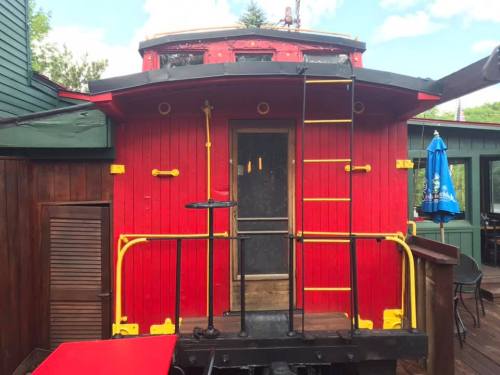 A bright red caboose with yellow accents, featuring a ladder and surrounded by greenery and outdoor seating.