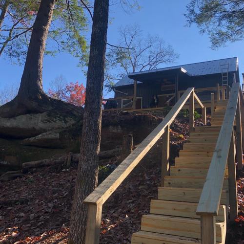 A wooden staircase leads up to a cabin surrounded by trees and autumn foliage. Clear blue sky above.