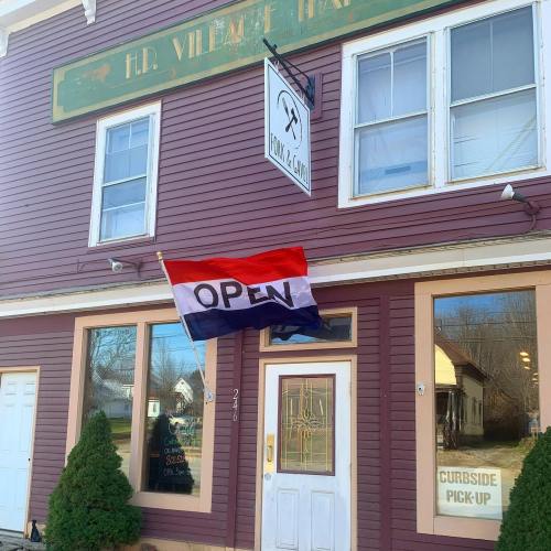 A storefront with a red, white, and blue "OPEN" flag hanging outside, surrounded by green shrubs.