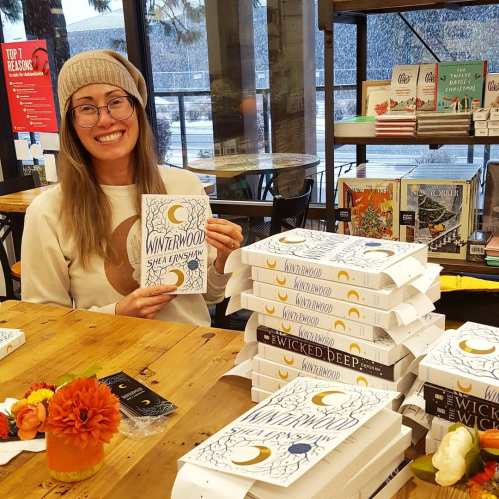 A smiling person holds a book titled "Winterwood" in a cozy bookstore with stacks of books and a snowy view outside.