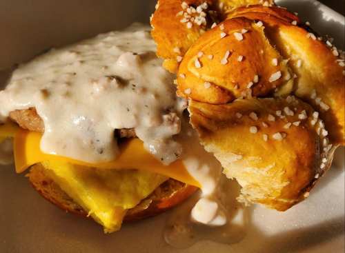 A close-up of a breakfast sandwich with sausage, cheese, and creamy gravy on a soft pretzel bun.