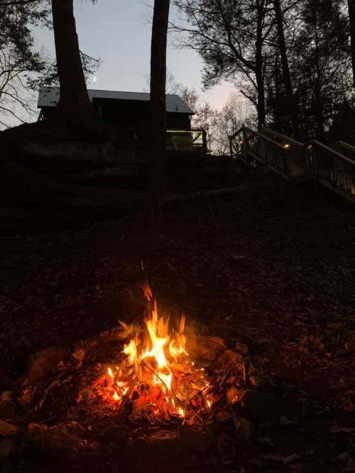 A glowing campfire in the foreground with a cabin and trees silhouetted against a twilight sky in the background.