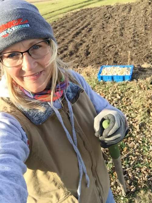 A smiling person in a beanie and gloves holds a shovel, standing in a field with a blue crate nearby.