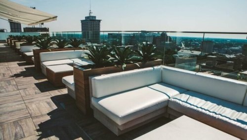 Rooftop lounge area with white seating and potted plants, overlooking a city skyline on a sunny day.