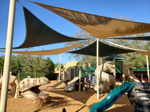 Playground with slides and climbing structures, shaded by large canopies, surrounded by trees and blue sky.
