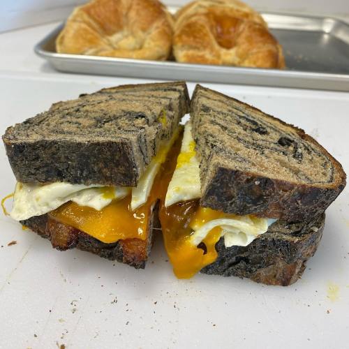 A sliced sandwich with black bread, melted cheese, fried egg, and bacon, with croissants in the background.