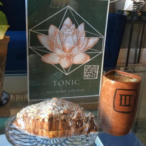 A pastry on a glass plate next to a ceramic cup, with a sign for "Tonic Alchemy Lounge" in the background.