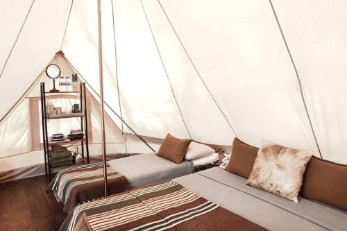 Cozy glamping tent interior with two beds, decorative pillows, and a shelf with toiletries and books.