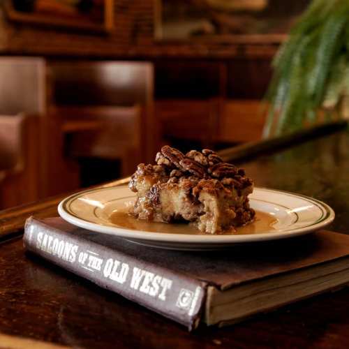 A slice of pecan pie on a plate, resting on a book titled "Saloons of the Old West," with a rustic background.