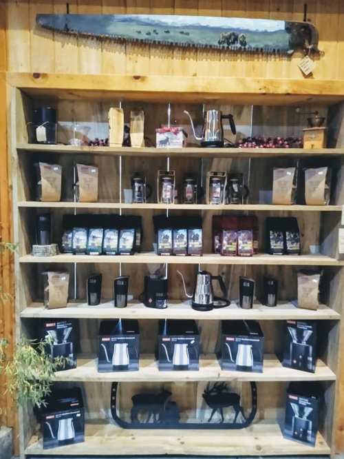 A wooden shelf displays various coffee products, including bags, mugs, and brewing equipment, with a saw hanging above.