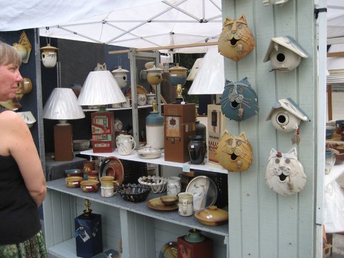 A woman stands near a market stall displaying various pottery items and whimsical animal-shaped birdhouses.