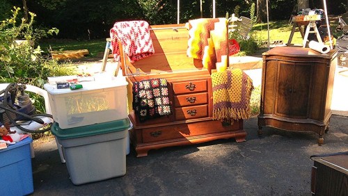 A collection of furniture and items, including a wooden chest, a small cabinet, and various blankets, in a yard sale setting.