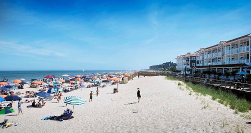A crowded beach with colorful umbrellas, people enjoying the sun, and a boardwalk alongside beachside buildings.