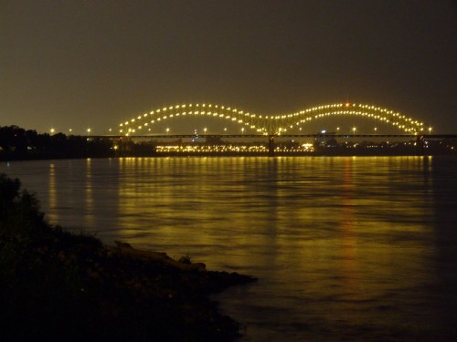 A brightly lit bridge spans a river at night, reflecting lights on the water's surface.