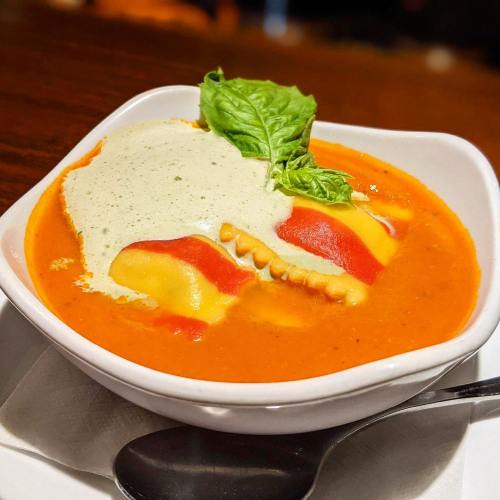 A bowl of vibrant tomato soup topped with colorful ravioli and a drizzle of creamy sauce, garnished with fresh basil.