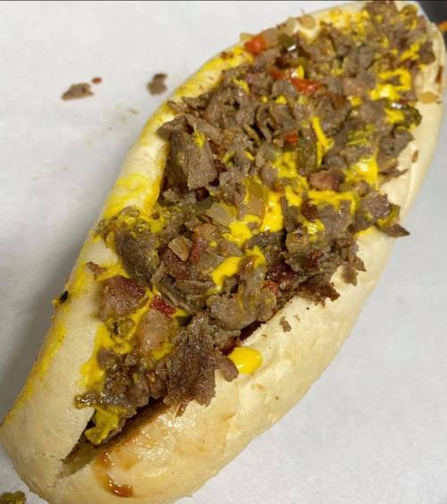 A loaded hot dog topped with chopped beef, mustard, and diced vegetables in a soft bun.