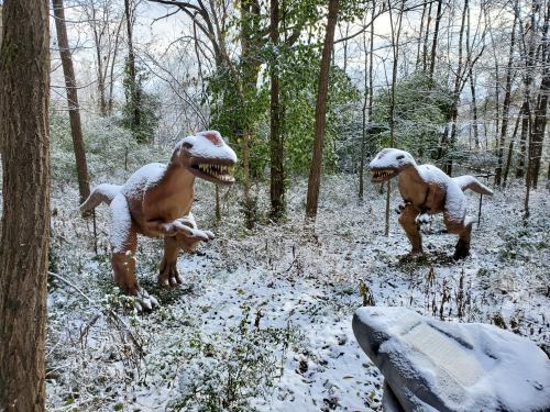 Two dinosaur sculptures covered in snow stand among trees in a snowy forest.