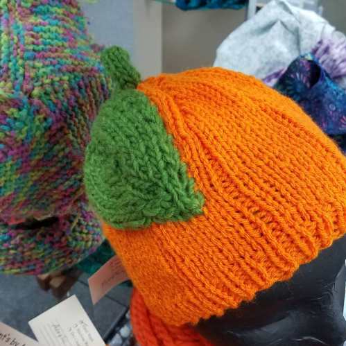 A vibrant orange knit hat with a green leaf detail on top, displayed on a mannequin head.