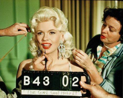 A glamorous woman with blonde hair and red lips poses for a film scene, while two people adjust her makeup and hair.