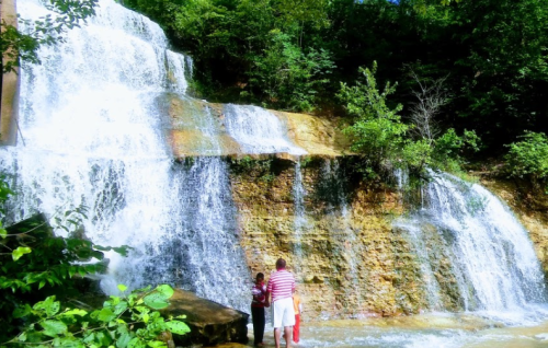A waterfall cascades over rocky terrain, with two people standing at its base surrounded by lush greenery.