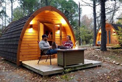 A couple sits by a fire pit outside cozy wooden cabins in a forested area, enjoying a peaceful evening.