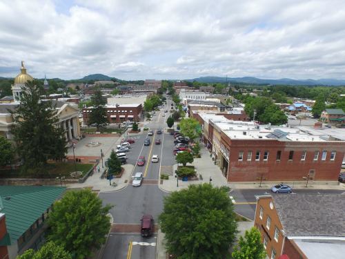 Aerial view of a small town with shops, trees, and mountains in the background under a cloudy sky.