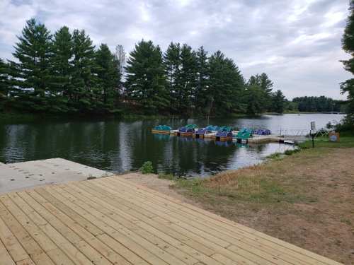 A serene lake scene with a wooden dock and colorful paddle boats, surrounded by lush green trees under a cloudy sky.