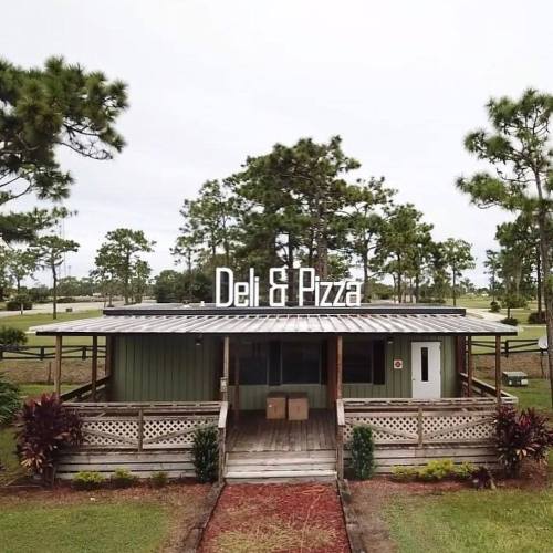 A small deli and pizza shop with a wooden deck, surrounded by trees and greenery. Sign reads "Deli & Pizza."