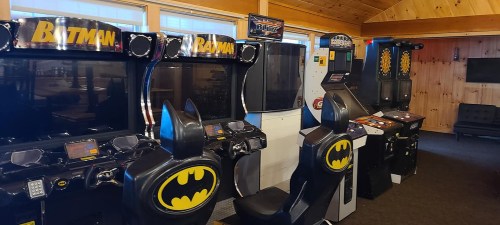 A row of arcade machines themed around Batman, featuring black seats with bat ears in a cozy wooden room.
