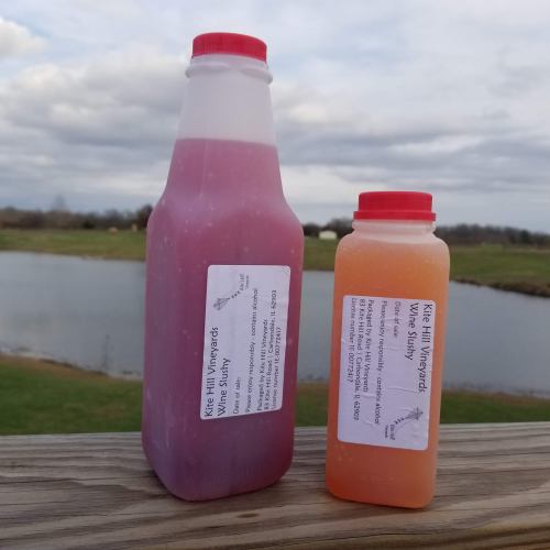 Two bottles of wine slushy from Kite Hill Vineyards, one pink and one orange, placed on a wooden railing by a lake.