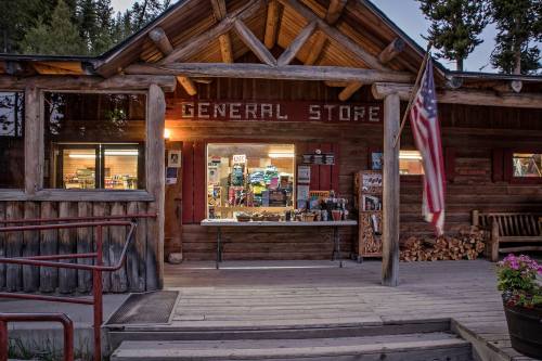 A rustic general store with a wooden facade, displaying goods outside and an American flag hanging nearby.