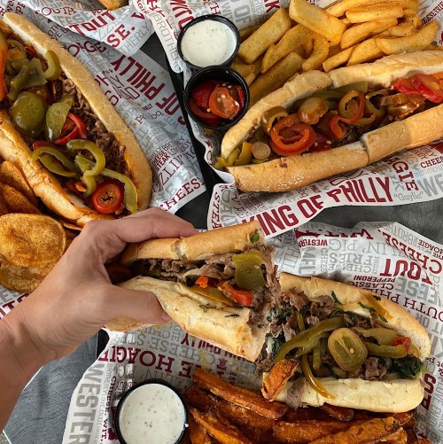 A hand holds a loaded sandwich with peppers, alongside fries and chips on a patterned paper background.