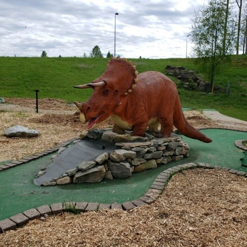 A large dinosaur sculpture, resembling a triceratops, stands on a rock formation in a grassy area.