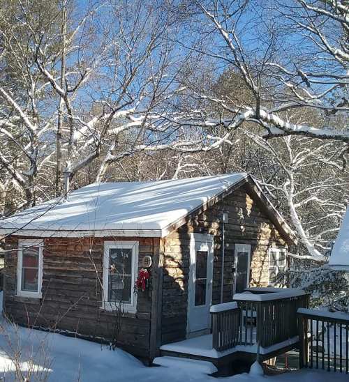 A cozy log cabin covered in snow, surrounded by trees with a clear blue sky above.
