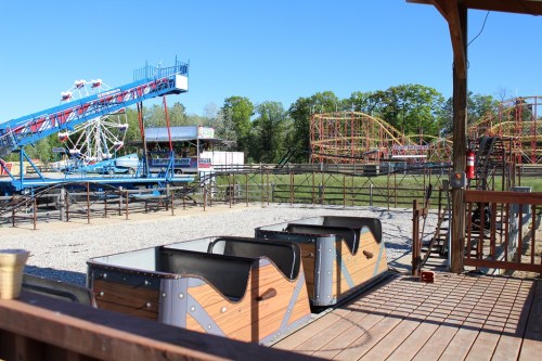 A view of an amusement park with a roller coaster, Ferris wheel, and empty ride cars under a clear blue sky.