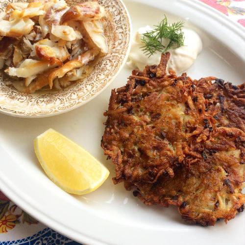 A plate featuring a crispy potato latke, a side of chopped fish, a lemon wedge, and a dollop of sour cream.