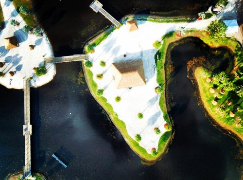 Aerial view of islands with sandy areas, huts, and surrounding water, featuring lush greenery and wooden walkways.