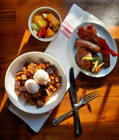 A hearty breakfast plate with potatoes, poached eggs, bacon, sausage, and a bowl of mixed fruit on a wooden table.